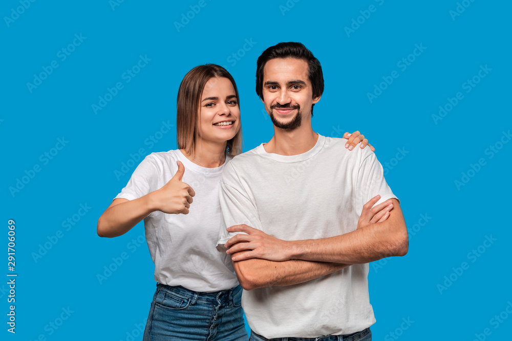 Happy couple of a young blond woman and brunet bearded man with mustaches in white t-shirts and blue jeans posing isolated over blue background. Concept of an ideal couple. Models holding thumbs up Happy couple of a young blond woman and brunet bearded man with mustaches in white t-shirts and blue jeans posing isolated over blue background. Concept of an ideal couple. Models holding thumbs up
