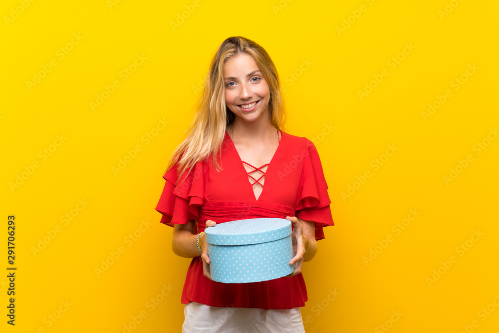 Blonde young woman over isolated yellow background holding gift box Blonde young woman over isolated yellow background holding gift box