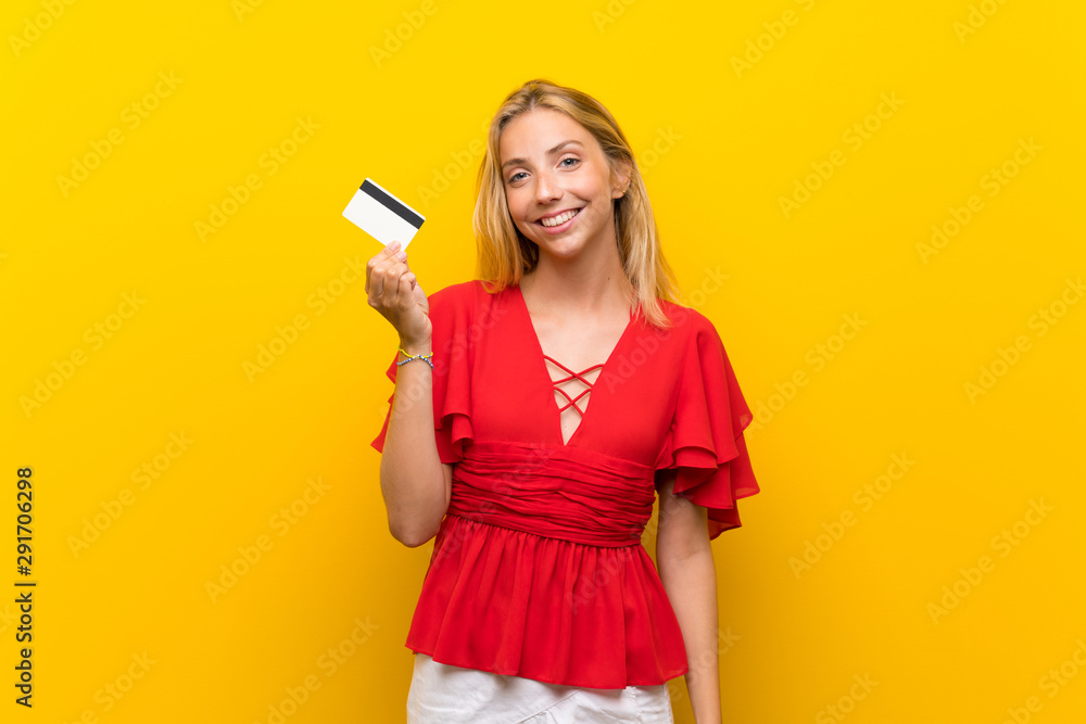 Blonde young woman over isolated yellow background holding a credit card Blonde young woman over isolated yellow background holding a credit card