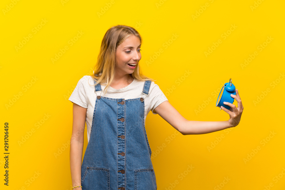 Blonde young woman over isolated yellow background holding vintage alarm clock Blonde young woman over isolated yellow background holding vintage alarm clock