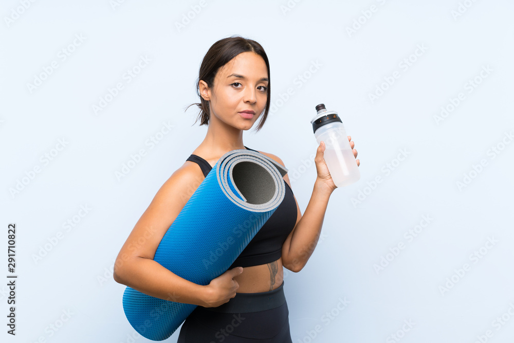Fototapeta premium Young sport girl with mat and with a bottle of water over isolated blue background