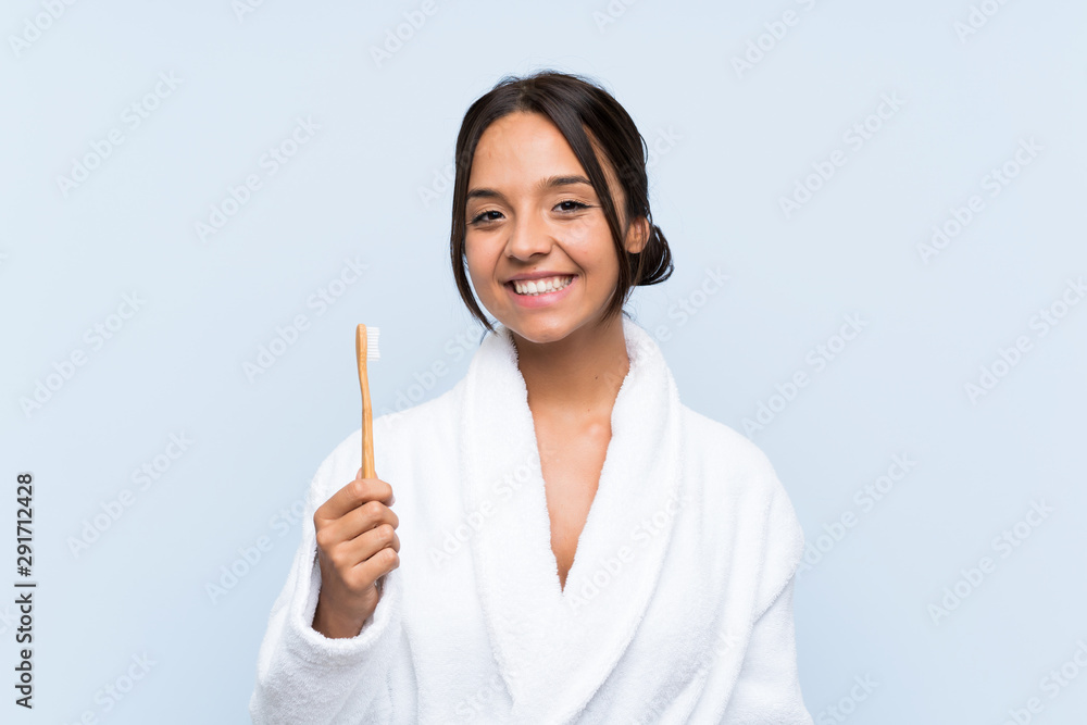 Young brunette woman in bathrobe brushing her teeth over isolated blue background smiling a lot Young brunette woman in bathrobe brushing her teeth over isolated blue background smiling a lot