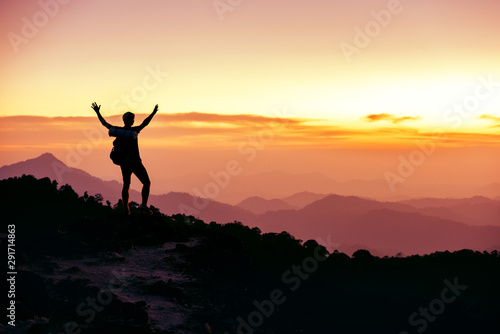 Hiker's silhouette stands on mountain top against sunset