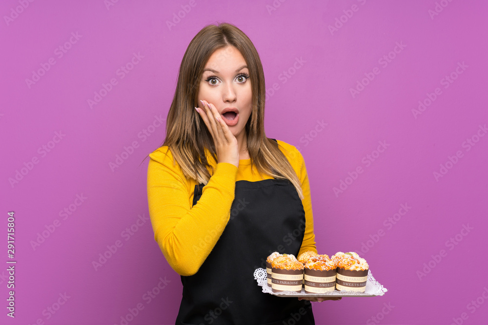 Teenager girl holding lots of different mini cakes over isolated purple background with surprise facial expression Teenager girl holding lots of different mini cakes over isolated purple background with surprise facial expression