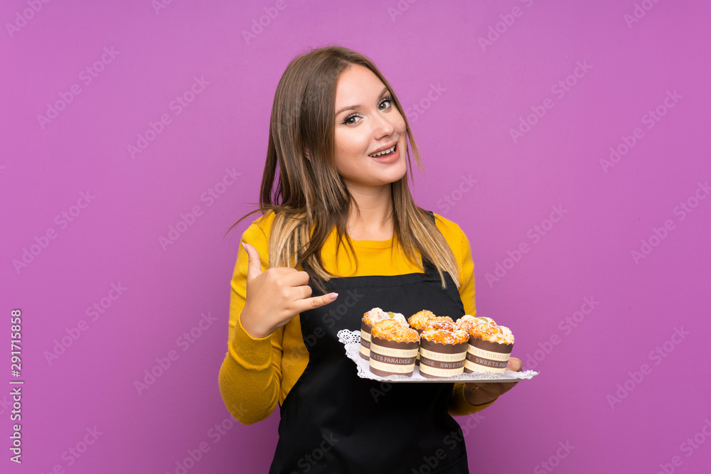 Teenager girl holding lots of different mini cakes over isolated purple background making phone gesture Teenager girl holding lots of different mini cakes over isolated purple background making phone gesture