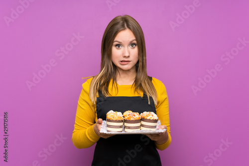 Teenager girl holding lots of different mini cakes over isolated purple background having doubts and with confuse face expression