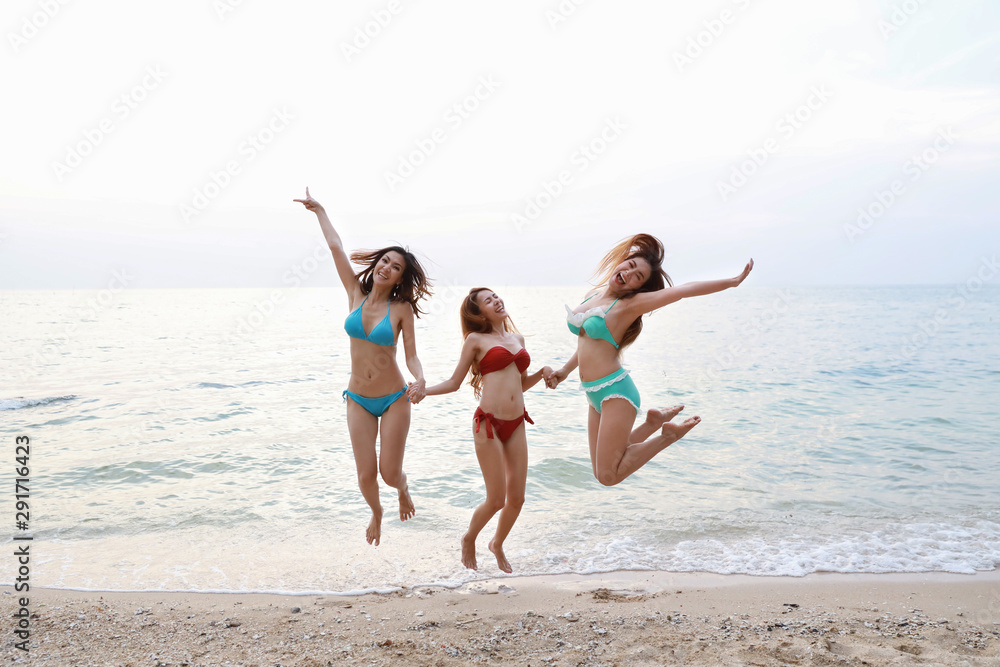 full shot of young asian beautiful and joyful girls in bikini who jumping and raising hands on tropical beach with happy and smile faces on holiday vacation with blue sky 