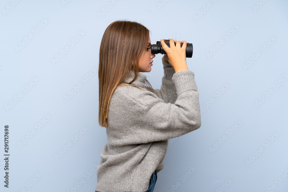 Teenager girl with sweater over isolated blue background with black binoculars Teenager girl with sweater over isolated blue background with black binoculars