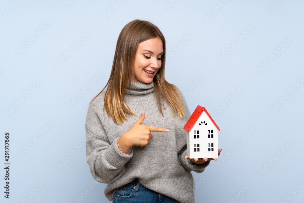 Teenager girl with sweater over isolated blue background holding a little house Teenager girl with sweater over isolated blue background holding a little house