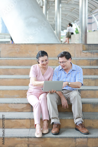 full length asian traveler couple husband and wife in pink and blue shirt with white nice hat sitting on the stairs in downtown and using computer to search location or shopping while taking a rest