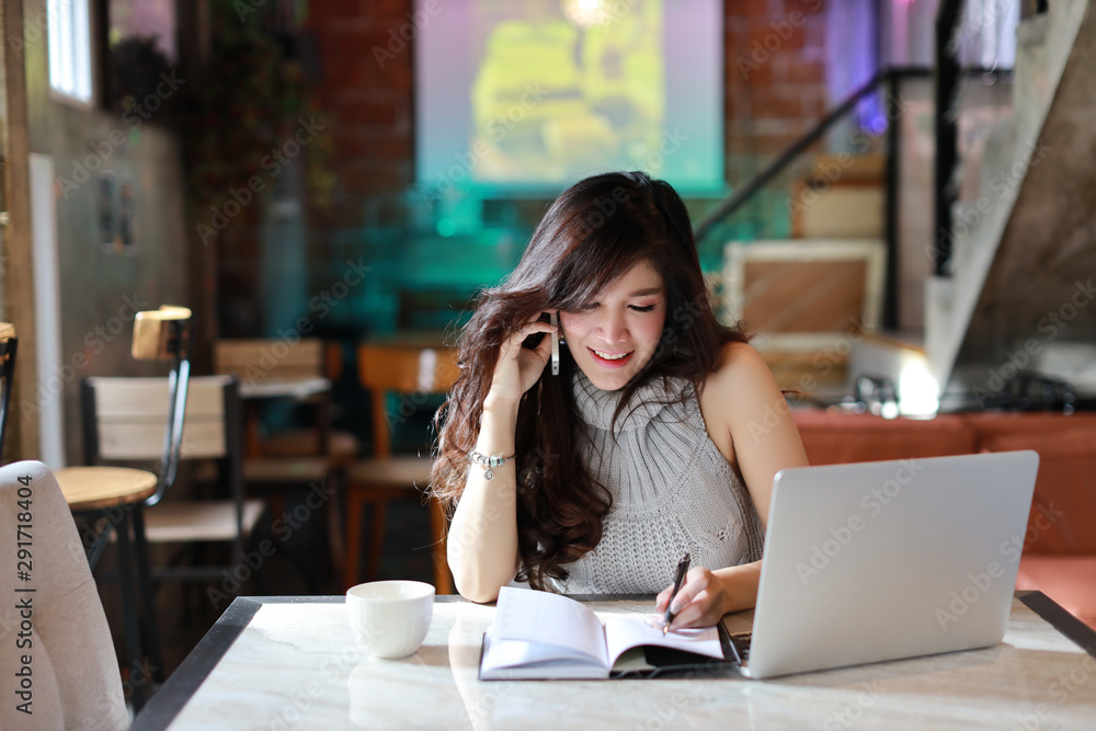 beautiful and attractive asian woman, long hair in grey casual dress working on paperwork while using cell phone and laptop computer in coffee shop beautiful and attractive asian woman, long hair in grey casual dress working on paperwork while using cell phone and laptop computer in coffee shop