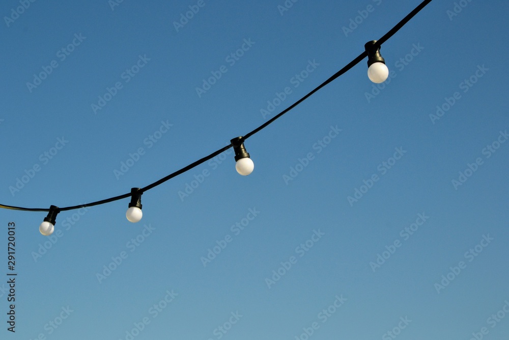 A string wire of outdoor light bulbs hanging against a clear blue sky. Light bulbs on the sky background. A string wire of outdoor light bulbs hanging against a clear blue sky. Light bulbs on the sky background.