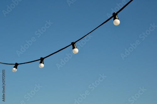 A string wire of outdoor light bulbs hanging against a clear blue sky. Light bulbs on the sky background. 