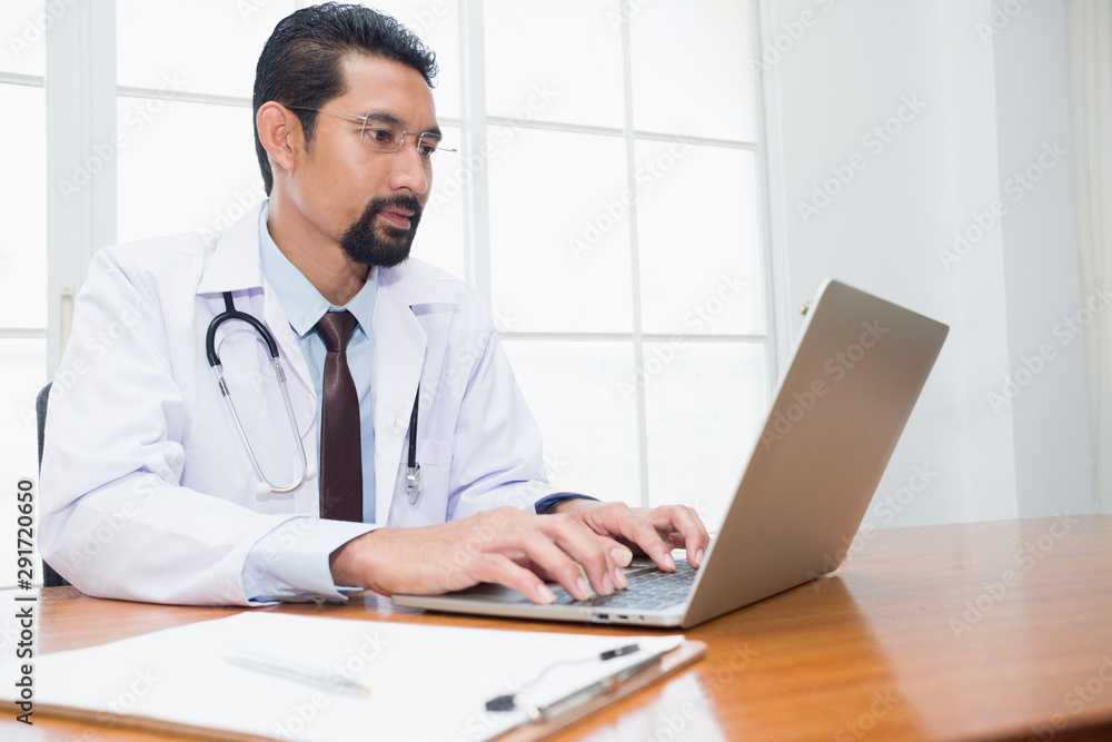 Adult Doctor wearing a medical robe with a stethoscope hanging on the neck while sitting working with a laptop in the workplace at the hospital. Adult Doctor wearing a medical robe with a stethoscope hanging on the neck while sitting working with a laptop in the workplace at the hospital.