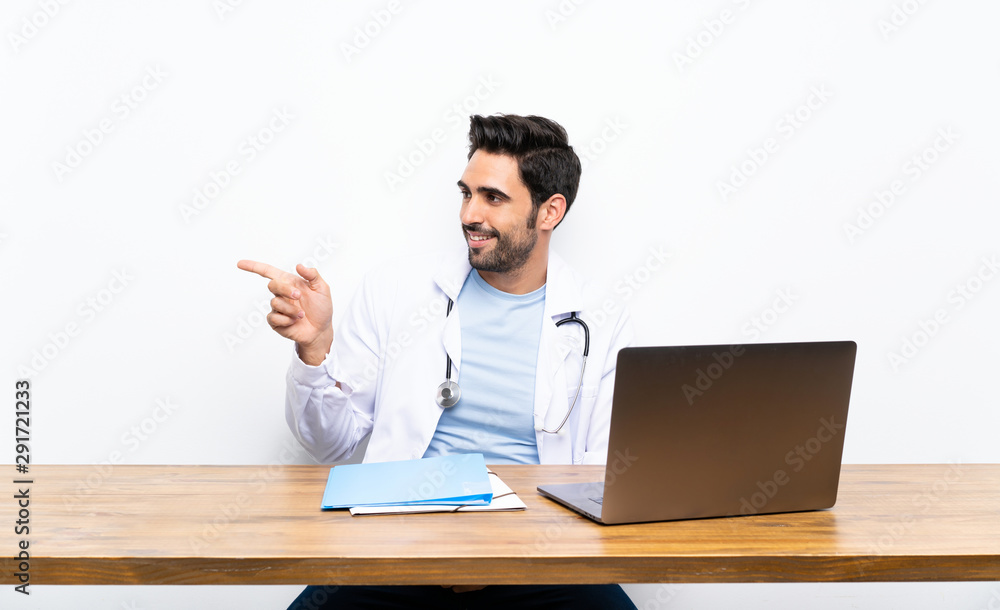 Young doctor man with his laptop over isolated wall pointing finger to the side Young doctor man with his laptop over isolated wall pointing finger to the side