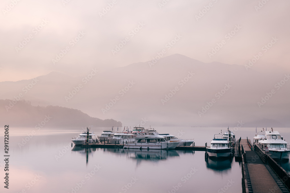 Fototapeta premium Beautiful tranquil landscape at Sun Moon lake in Nantao, Taiwan. Pier with boats and background of foggy mountains. Concept of peaceful, traanquility of nature.
