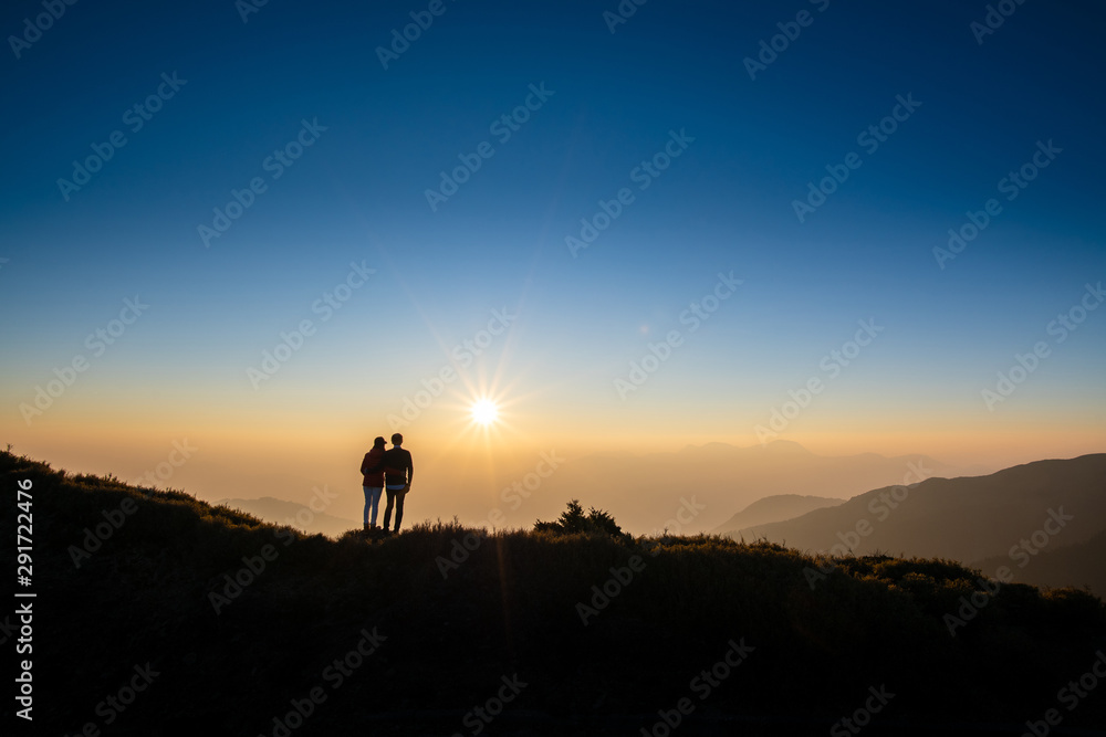 Silhoutte of a couple looking at sunset on top of hill with background of mountain and golden color of sunshine. Concept of romantic, forever love.