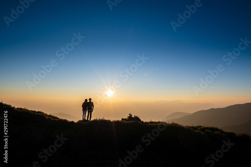 Silhoutte of a couple looking at sunset on top of hill with background of mountain and golden color of sunshine. Concept of romantic, forever love. © 9mot