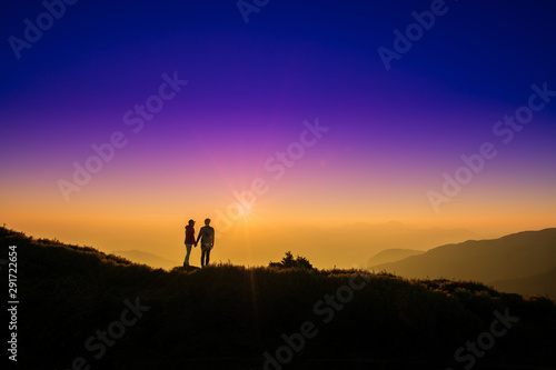 Silhoutte of a couple hold hand of each other looking at sunset on top of hill with background of mountain and colorful color of sunset. Concept of teamwork, successful.