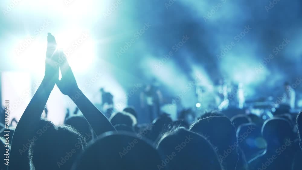 Back view of crowded concert hall with music fans applauding to favourite band. Shot against bright stage lights Back view of crowded concert hall with music fans applauding to favourite band. Shot against bright stage lights