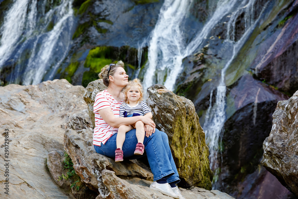 Cute toddler girl and mother sitting near water cascade of Powerscourt Waterfall, the highest waterfall in Ireland in co. Wicklow. Family time vacations with small children. Woman and baby child