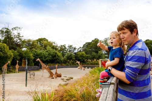 Canvas Print Cute adorable toddler girl and father watching and feeding giraffe in zoo