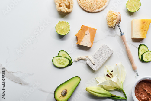 top view of natural soap pieces, body brush, loofah and pumice stone near fresh flower, lime, cucumber and avocado on marble surface