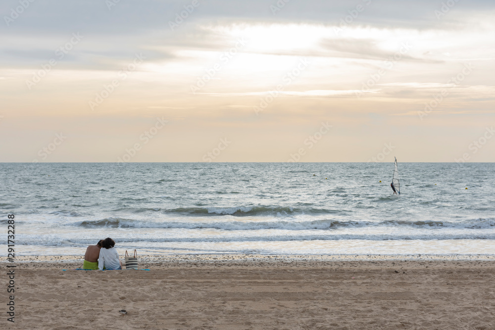 Dos personas observan el paisaje, sobre la arena de la playa, una persona practica  windsurf 