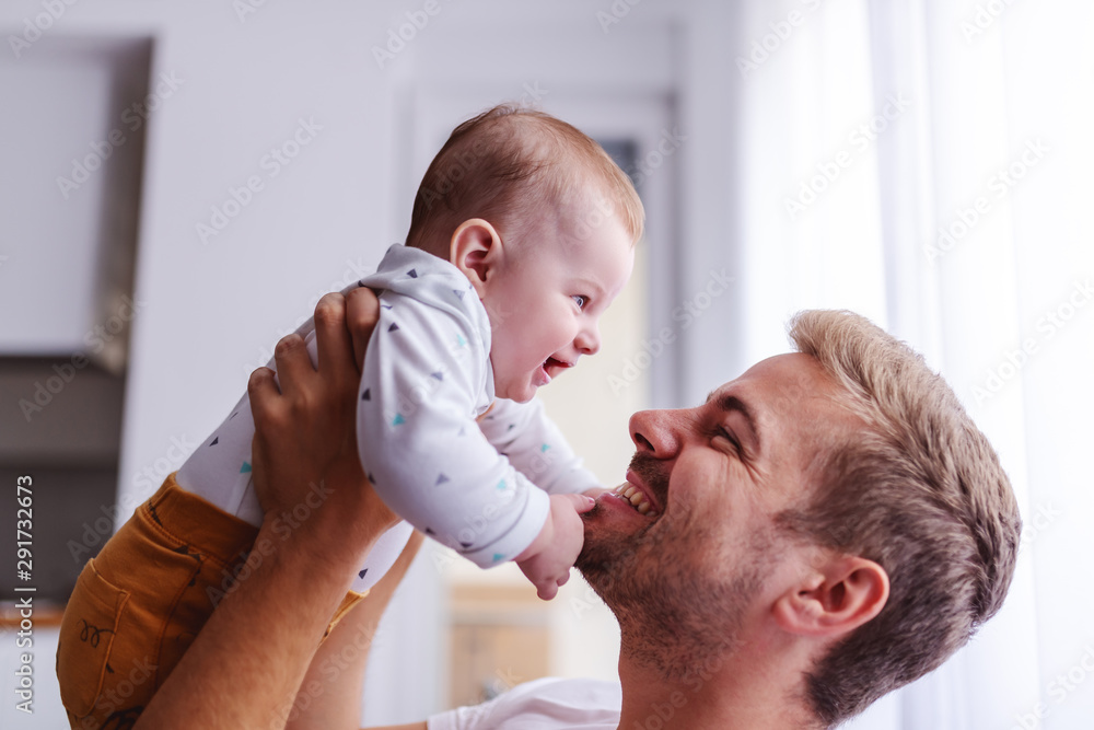 Happy smiling handsome young caucasian dad lifting his loving 6 months old son  while standing in living room next to window. Baby is touching dad's face  and laughing. Unconditional love concept. Photos