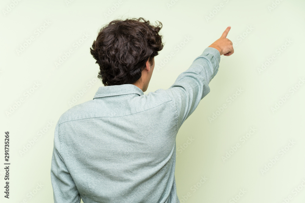 Young man over isolated green wall pointing back with the index finger Young man over isolated green wall pointing back with the index finger