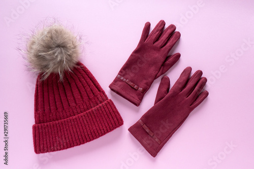 Cozy and warm winter flat lay with copy space. Dark red knitted hat with fur pompom and burgundy gloves on pink background