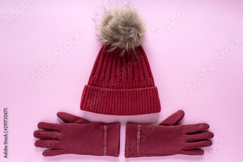 Cozy and warm winter flat lay with copy space. Dark red knitted hat with fur pompom and burgundy gloves on pink background