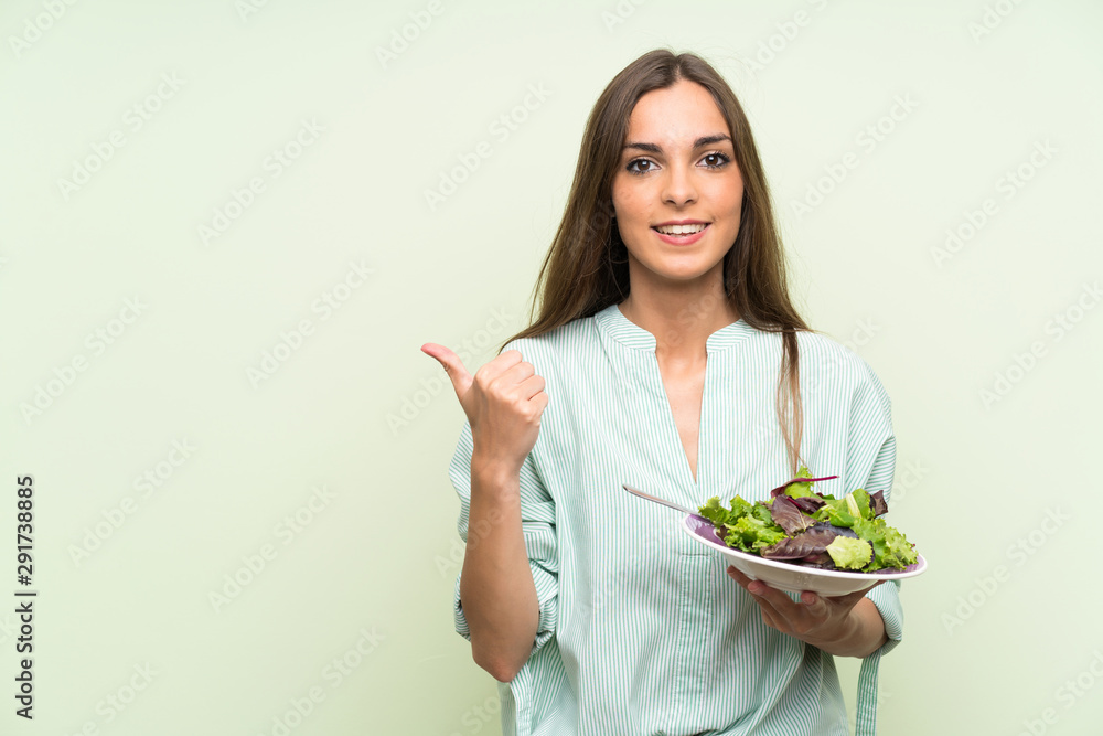 Young woman with salad over isolated green wall pointing to the side to present a product Young woman with salad over isolated green wall pointing to the side to present a product