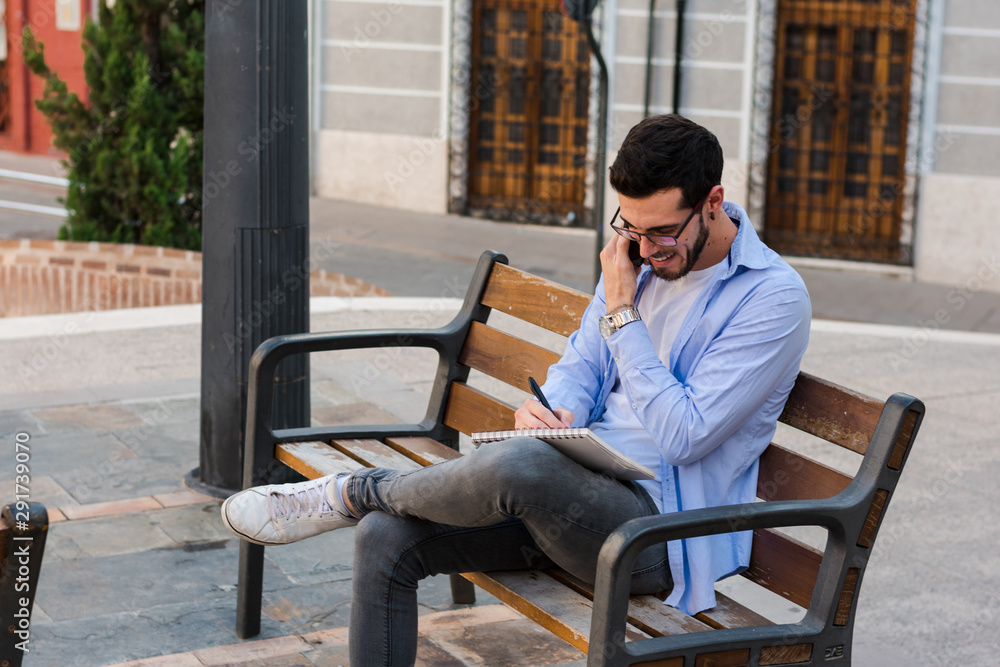 Young businessman is sitting on a bench while he is talking on the mobile and is writing in his notebook Young businessman is sitting on a bench while he is talking on the mobile and is writing in his notebook