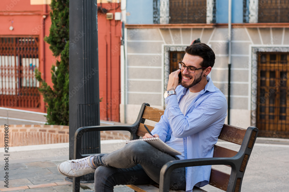 Young businessman is sitting on a bench while he is talking on the mobile and is writing in his notebook Young businessman is sitting on a bench while he is talking on the mobile and is writing in his notebook