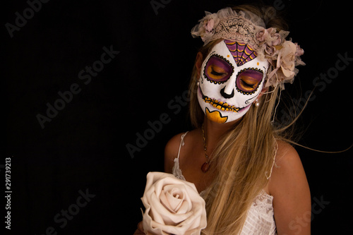 Young blonde and pretty girl  wearing makeup with Mexican Halloween makeup. She is posing in a white dress  a wreath and a flower in her hand