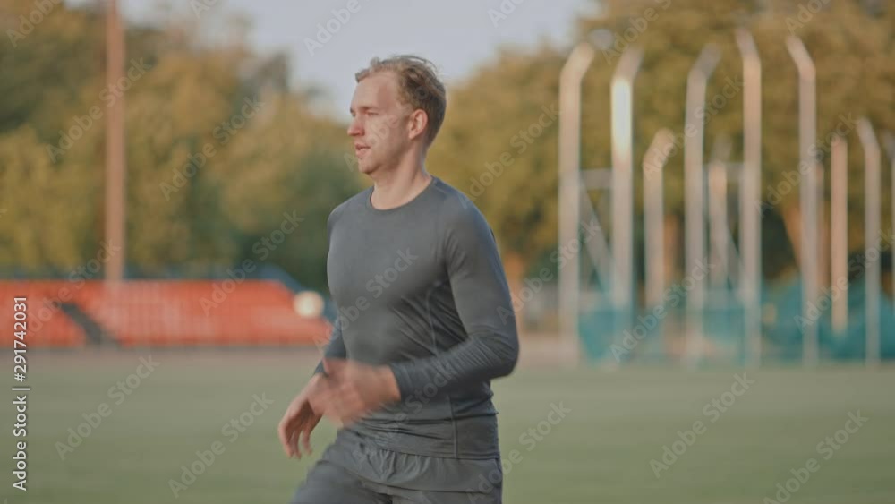 Athletic Fit Man in Grey Shirt and Shorts Jogging in the Stadium. He is Running Fast on a Warm Summer Afternoon. Athlete Doing His Routine Sports Practice. Slow Motion Tracking Shot. Athletic Fit Man in Grey Shirt and Shorts Jogging in the Stadium. He is Running Fast on a Warm Summer Afternoon. Athlete Doing His Routine Sports Practice. Slow Motion Tracking Shot.