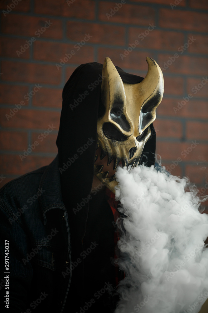 Vape man in devil mask and a hood smoking an electronic cigarette indoors.  Puffs of steam. Bad habit that is harmful to health. Vaping activity. Stock  Photo | Adobe Stock