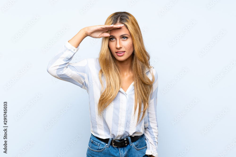 Young blonde woman over isolated blue background looking far away with hand to look something Young blonde woman over isolated blue background looking far away with hand to look something