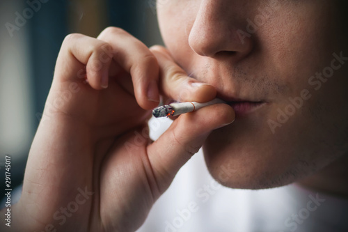 A young man with a stubble on his face inhales the smoke of a smoldering cigarette, holding it between his fingers.