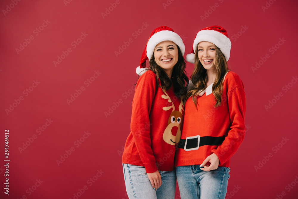 attractive and smiling women in sweaters and santa hats looking at camera isolated on red attractive and smiling women in sweaters and santa hats looking at camera isolated on red