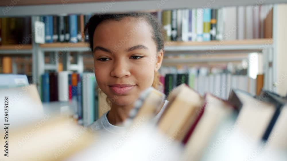 Portrait of cheerful African American teenager choosing books in school library touching textbooks smiling. Education, people and youth culture concept. Portrait of cheerful African American teenager choosing books in school library touching textbooks smiling. Education, people and youth culture concept.
