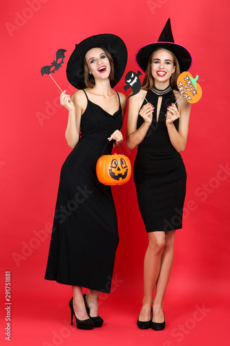Two young women in halloween costumes with pumpkin bucket and paper ghost and bat on red background