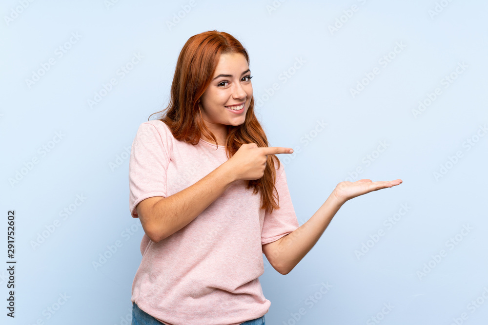 Teenager redhead girl over isolated blue background holding copyspace imaginary on the palm to insert an ad Teenager redhead girl over isolated blue background holding copyspace imaginary on the palm to insert an ad