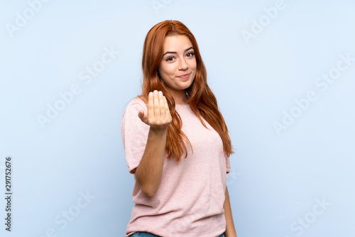 Teenager redhead girl over isolated blue background inviting to come with hand. Happy that you came