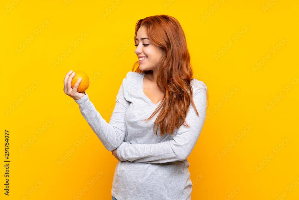 Teenager redhead girl holding an orange over isolated yellow background Teenager redhead girl holding an orange over isolated yellow background