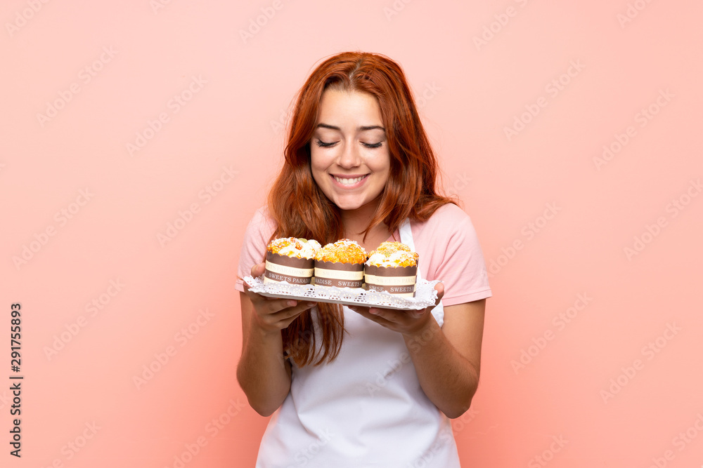 Teenager redhead girl holding lots of different mini cakes over isolated pink background Teenager redhead girl holding lots of different mini cakes over isolated pink background