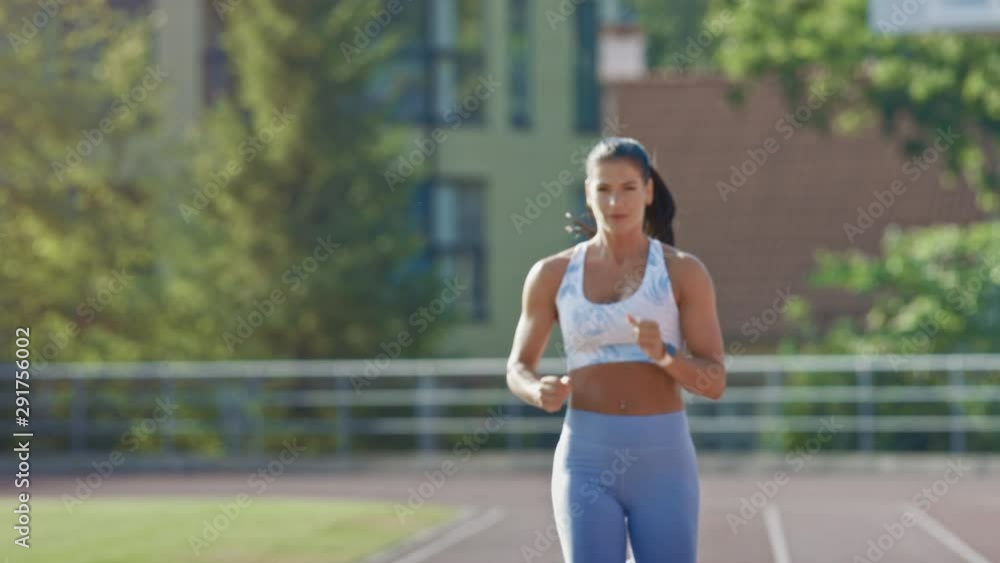 Happy and Smiling Fitness Woman in Light Blue Athletic Top and Leggings Jogging in a Stadium. She is Running on a Warm Summer Afternoon. Athlete Doing Her Routine Sports Practice on a Track. Happy and Smiling Fitness Woman in Light Blue Athletic Top and Leggings Jogging in a Stadium. She is Running on a Warm Summer Afternoon. Athlete Doing Her Routine Sports Practice on a Track.