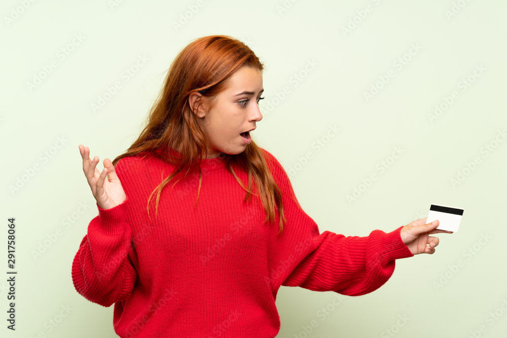 Teenager redhead girl with sweater over isolated green background holding a credit card Teenager redhead girl with sweater over isolated green background holding a credit card