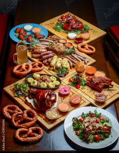 Flat-lay of Oktoberfest dinner table with grilled meat sausages, pretzel pastry, potatoes, cucumber salad, sauces, beers